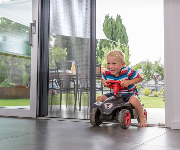 Kleinkind fährt auf einem Bobbycar durch eine Insektenschutztür ins Innere.